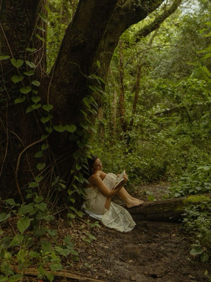 a woman sitting under a tree in the forest reading a book while wearing a white dress