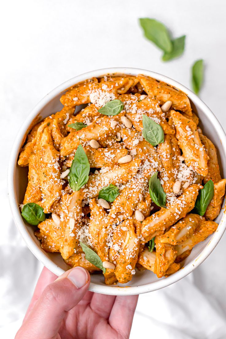 a hand holding a bowl of pasta with basil and pine nuts