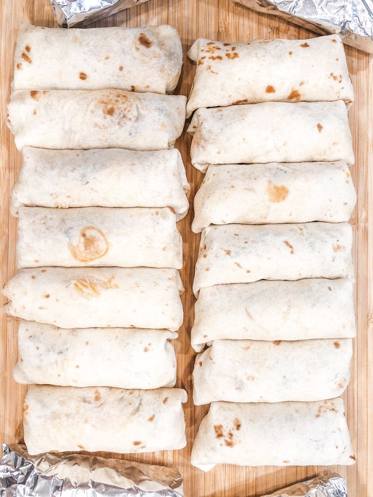 six burritos on a wooden cutting board lined with aluminum foil, ready to be baked