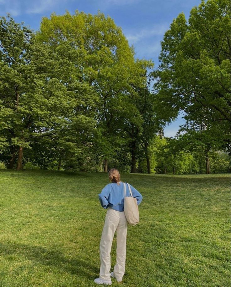 a woman standing in the grass with her back to the camera and looking at trees