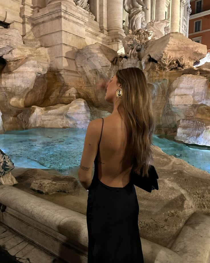 a woman in a black dress standing next to a fountain with statues on the sides