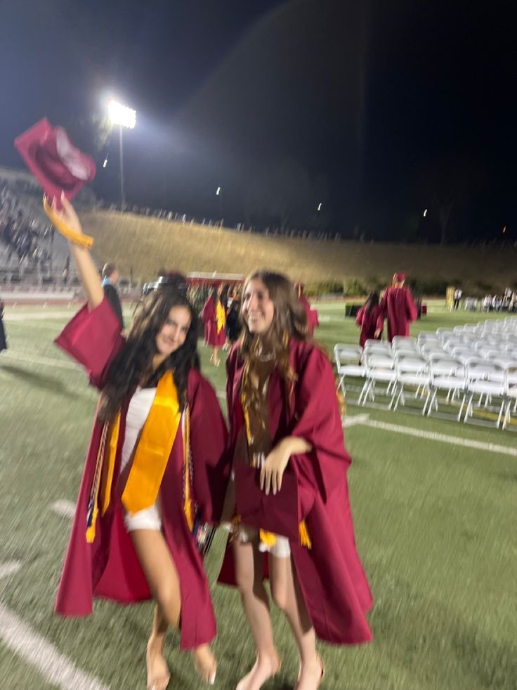 two young women in graduation gowns and caps are dancing on the field at night