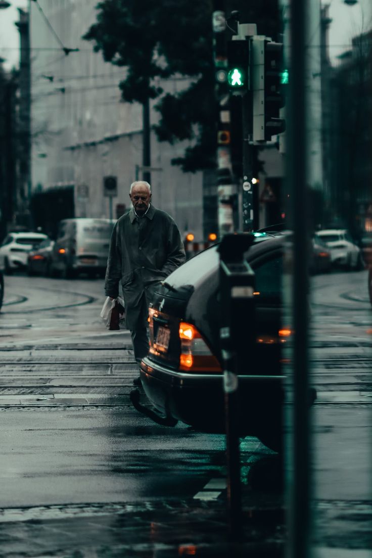 a man walking across a street next to a car on a rain soaked road with traffic lights