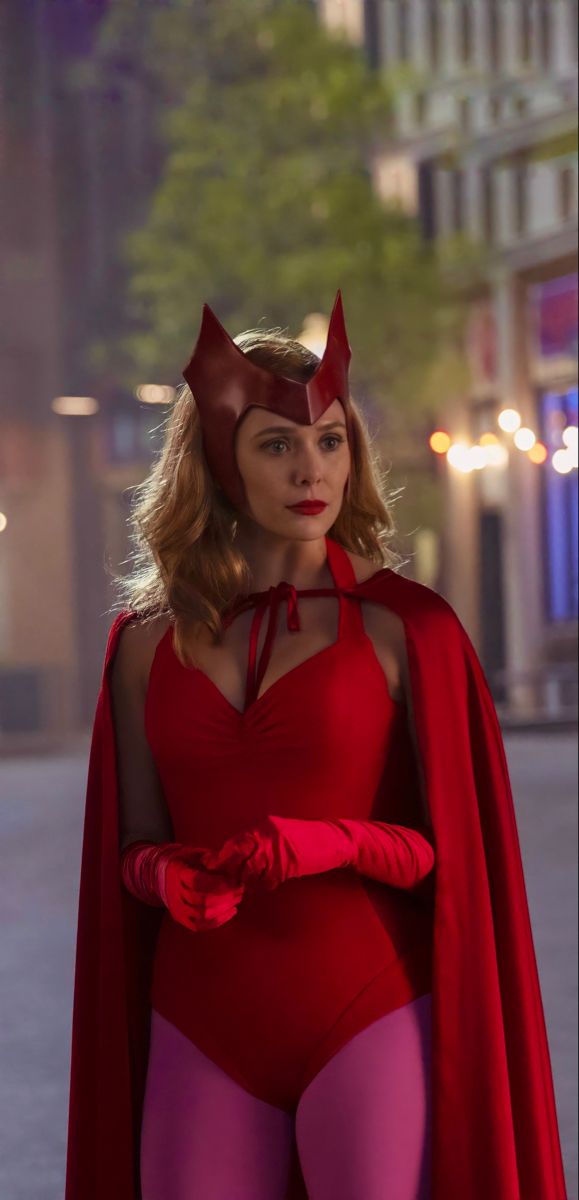 a woman in a red costume and cape standing on the street with her arms crossed