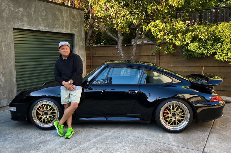 a man standing next to a black sports car in front of a garage with his feet on the ground