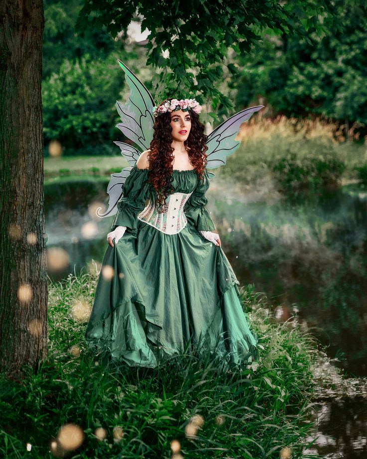 a woman dressed as a fairy standing in the grass next to a tree and water