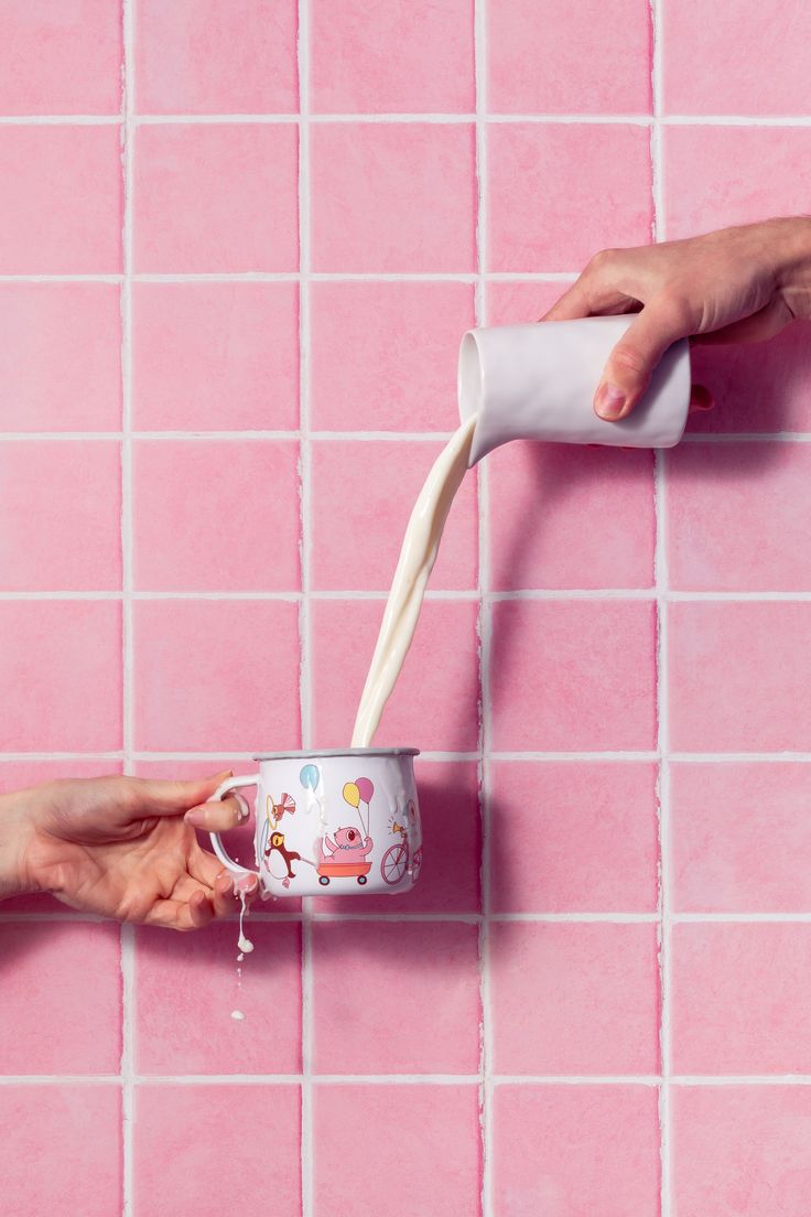 someone is pouring milk into a can on a pink tile wall with two hands reaching for it