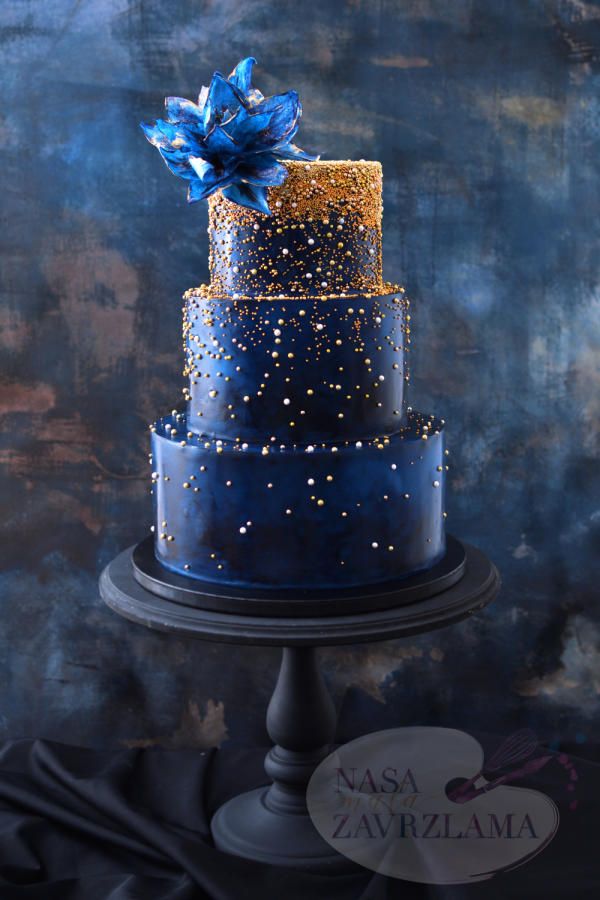 a three tiered blue and gold wedding cake on a black stand against a dark background