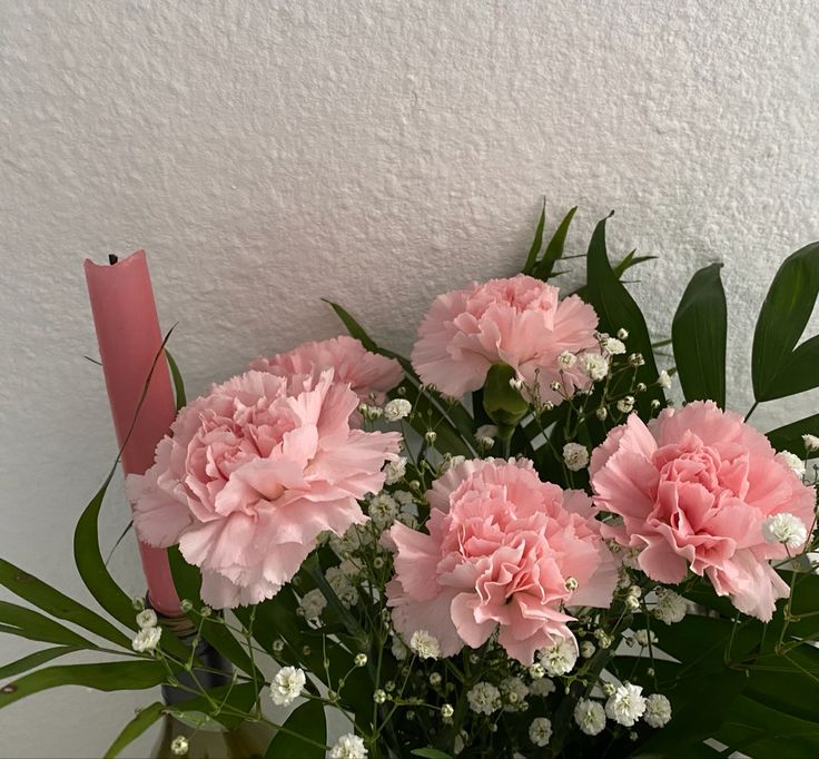 pink carnations in a vase with greenery and a candle on the wall