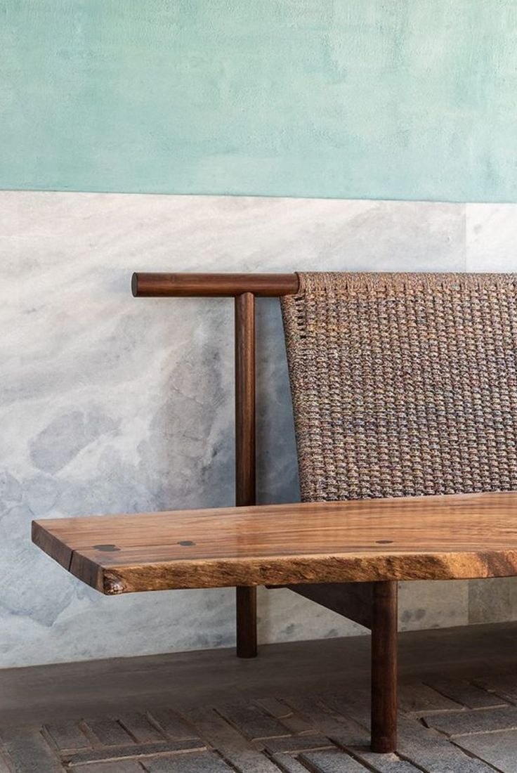 a wooden bench sitting in front of a wall next to a brick floor with a woven seat on it