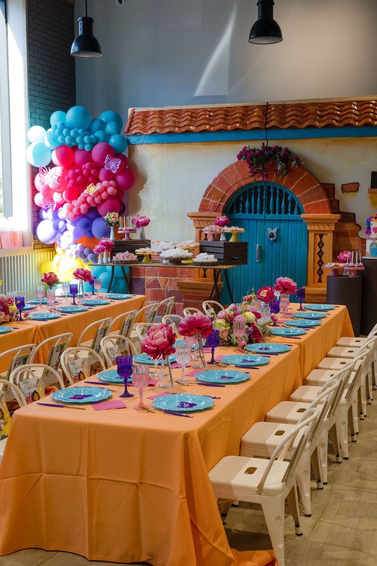 A long table set with orange and blue plates, vibrant floral centerpieces, and colorful balloon garlands, capturing the fun and magical vibe of the Encanto party. Colorful Birthday Decorations, Encanto Birthday Party Ideas, I Do Party, Encanto Theme, Birthday Setup, Encanto Birthday Party, Colorful Table Setting, Encanto Party, Colorful Birthday