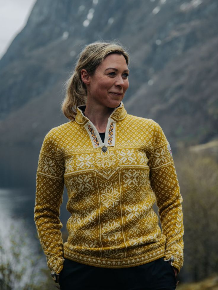 a woman wearing a yellow sweater standing in front of a body of water with mountains in the background