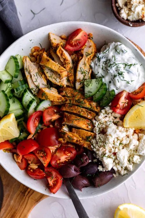 a white bowl filled with chicken, cucumber, tomatoes and other food items