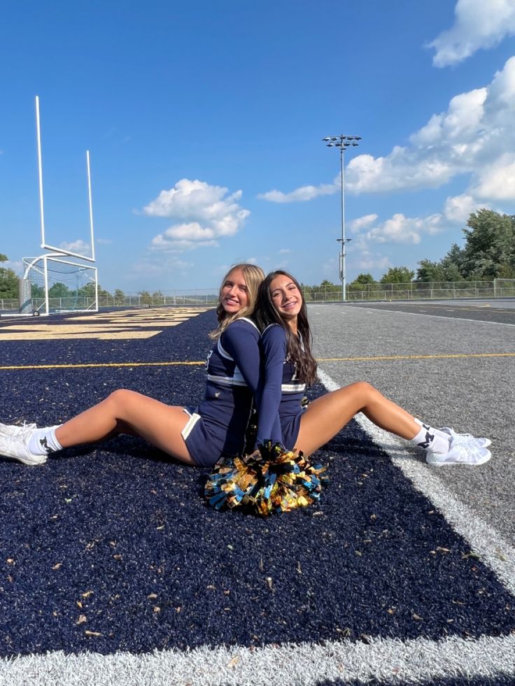 two cheerleaders sitting on the side of an empty parking lot smiling at the camera