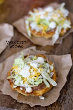 two mexican food items sitting on top of brown parchment paper with shredded cheese and lettuce