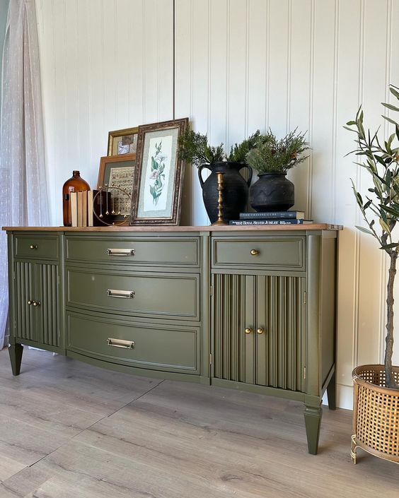 an olive green buffet cabinet with plants and pictures on top, in front of a white paneled wall