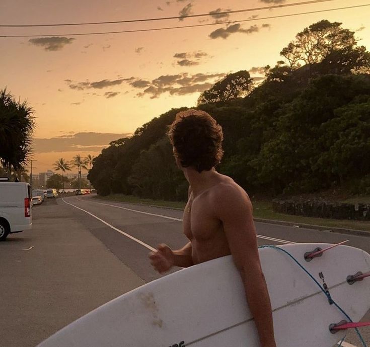 a shirtless man holding a surfboard on the side of the road at sunset