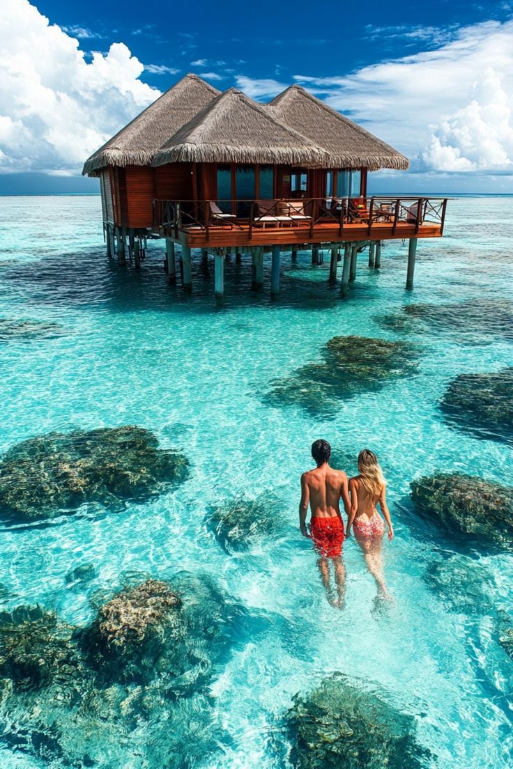 two people are walking in the water near a house on stilts over clear blue water