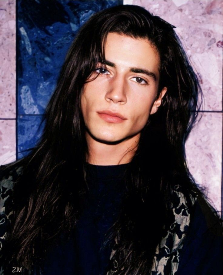 a young man with long hair and blue eyes posing for a photo in front of a tile wall