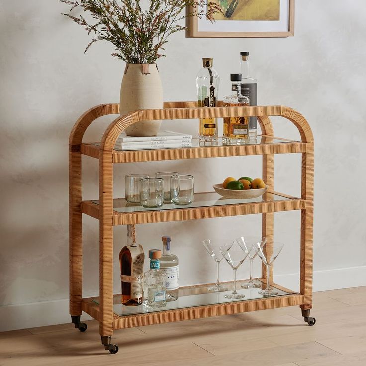 a bamboo bar cart with drinks and glasses on the top, in front of a painting