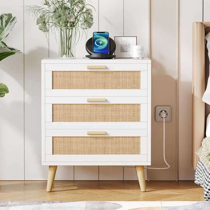 a white dresser with wicker drawers next to a plant and phone charger on top