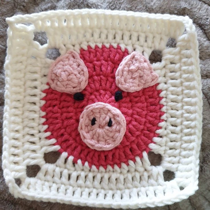 a crocheted square with a pig face on the front and bottom, sitting on a carpet