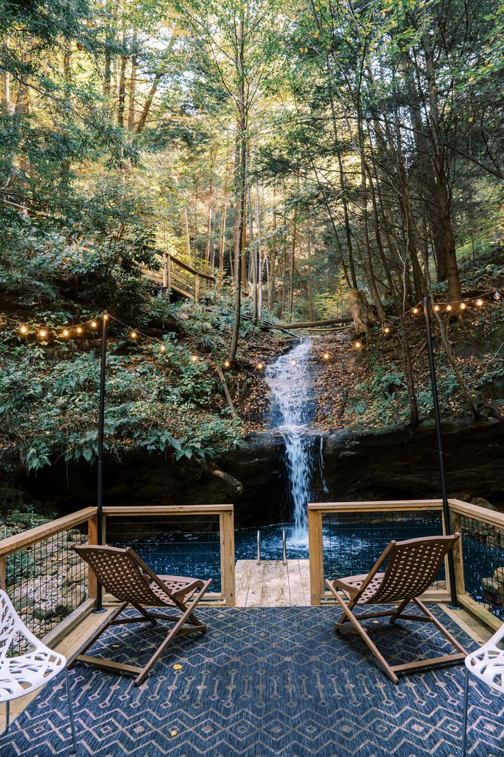 two lawn chairs sitting on top of a wooden deck next to a waterfall in the woods