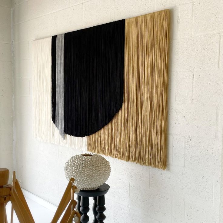 a wall hanging with black, white and tan colors on it next to a wooden chair