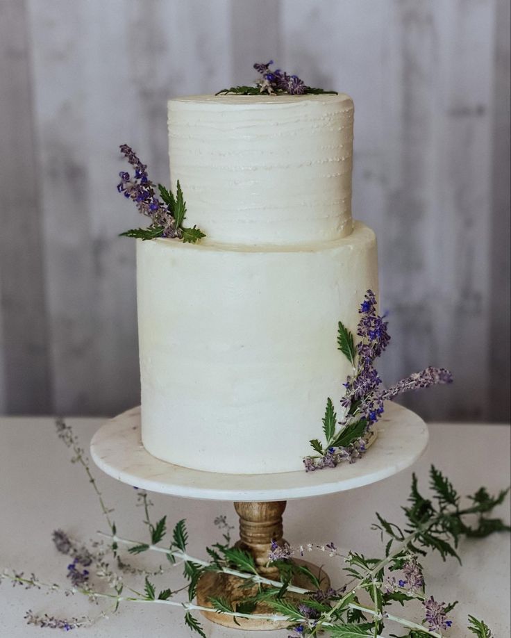 a three tiered white wedding cake with purple flowers on top and greenery around it