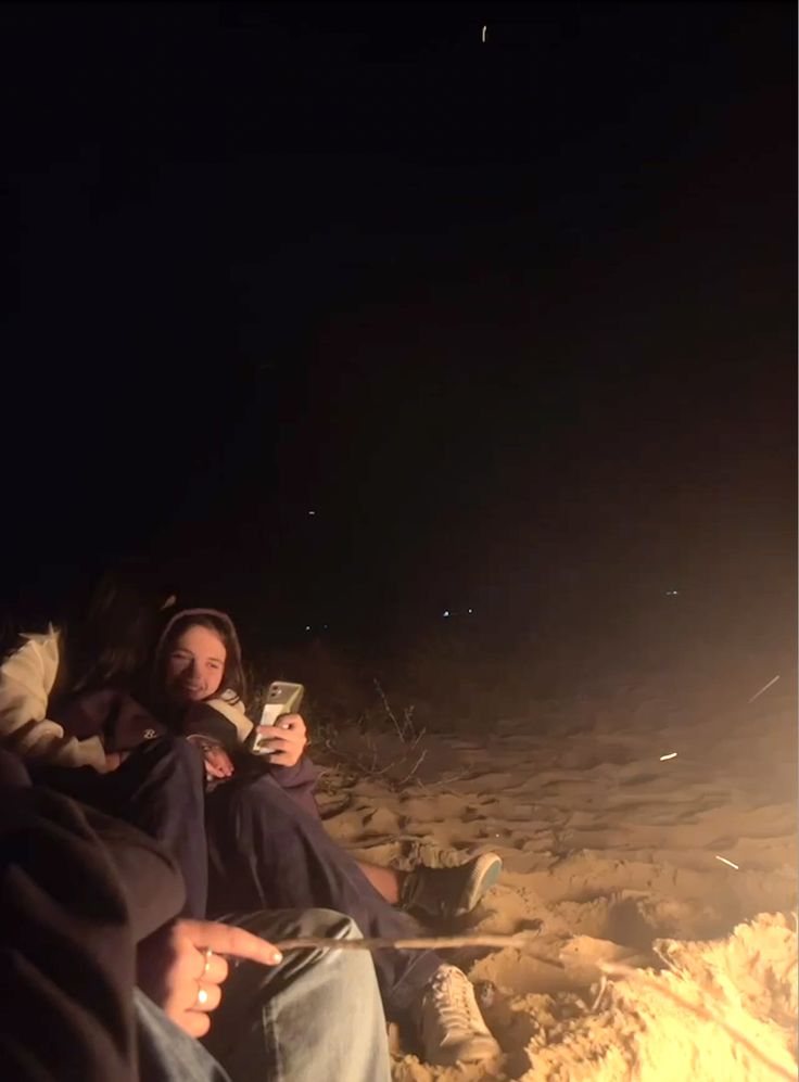 two people sitting next to each other on the beach at night with their cell phones