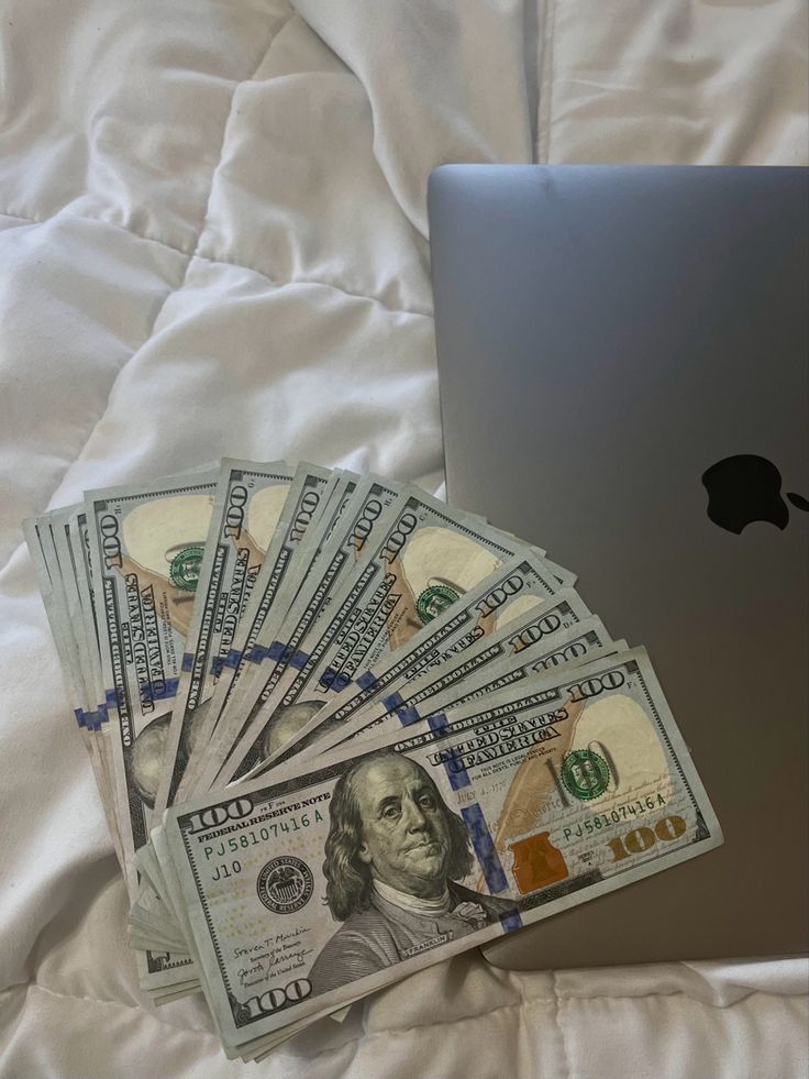 a pile of money sitting next to an apple laptop on top of a white bed