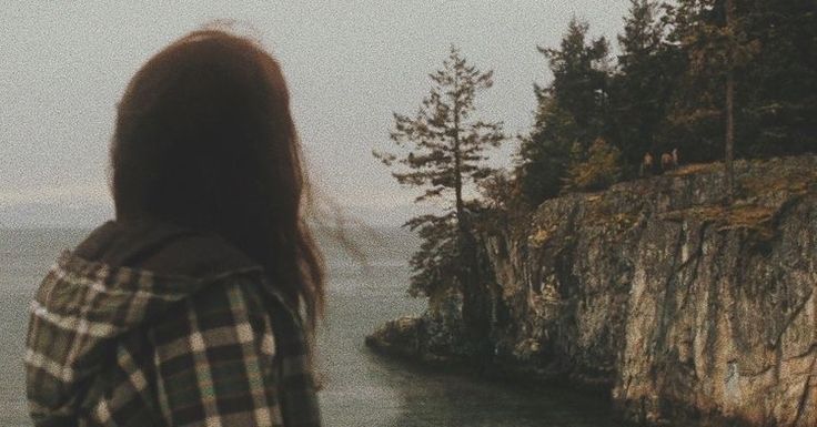 a woman standing on the edge of a cliff looking out to sea with trees in the background