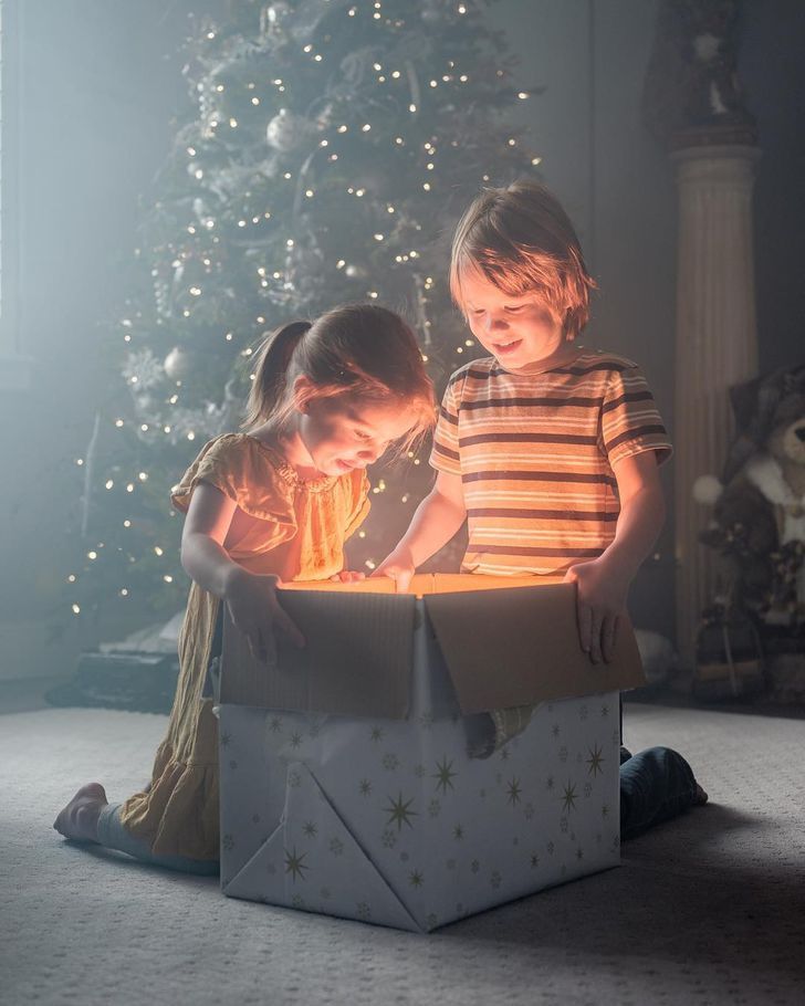 two young children playing with a lit candle in a gift box next to a christmas tree