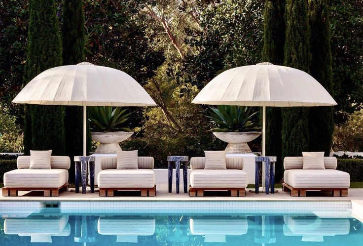 two lounge chairs with umbrellas next to a swimming pool in front of some trees