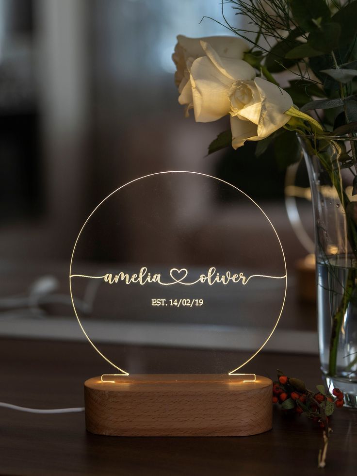 a glass vase with flowers and a personalized acrylic sign on it next to a wooden stand