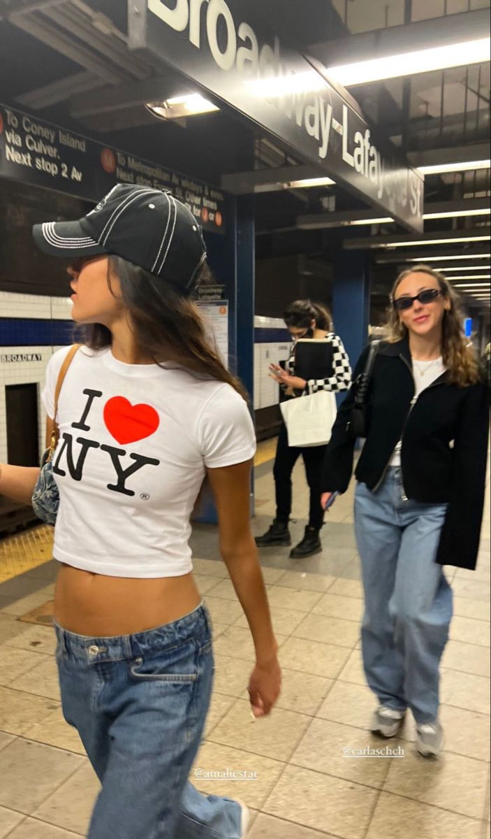 two women in jeans and baseball caps are walking through the subway station, one is wearing a t - shirt that says i love ny