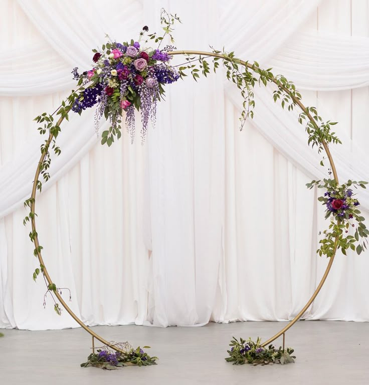 a circular floral arrangement with purple flowers and greenery in front of a white backdrop