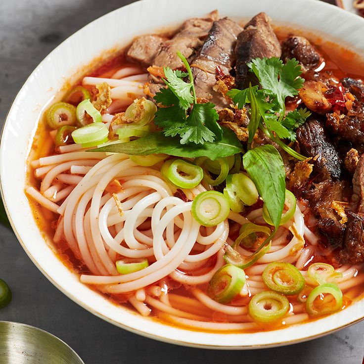 a white bowl filled with noodles and meat on top of a table next to green peppers