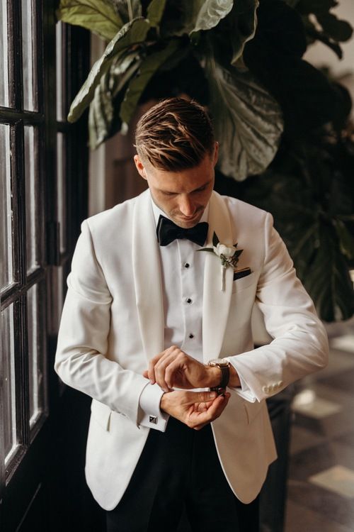 a man in a white tuxedo looking down at his watch while standing next to a window