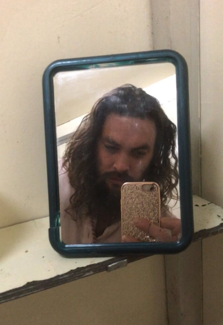 a man with long hair is looking at his cell phone in the mirror while brushing his teeth