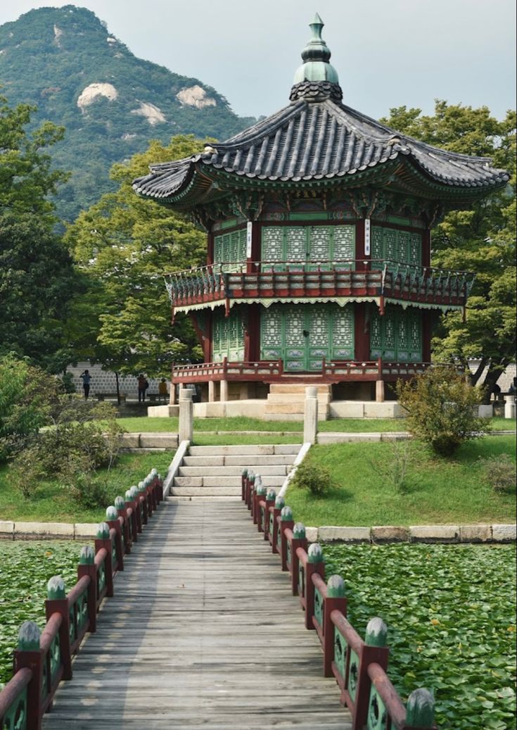 a wooden walkway leading to a pagoda in the middle of a park with mountains in the background