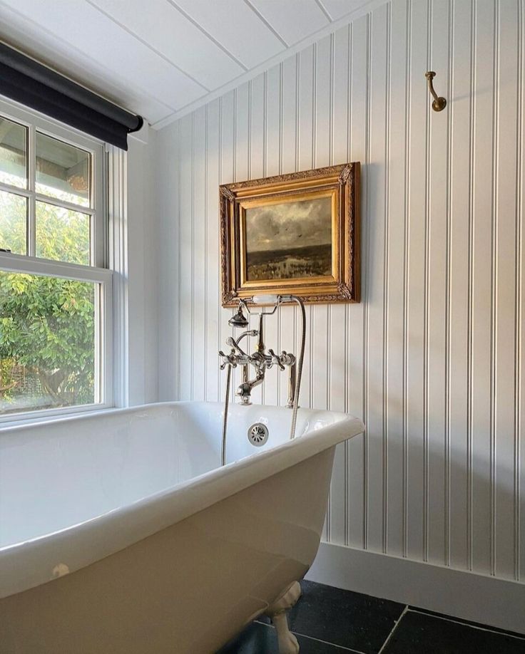 an old fashioned bathtub in front of a window with a painting on the wall