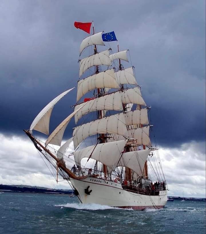 a large white sail boat sailing in the ocean under a cloudy sky with red, white and blue flags