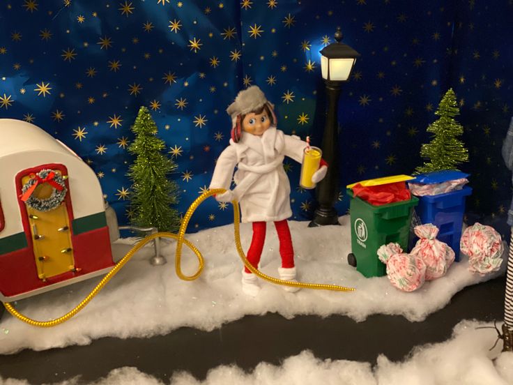 a doll is holding a hose next to a toy trailer and christmas trees in the snow