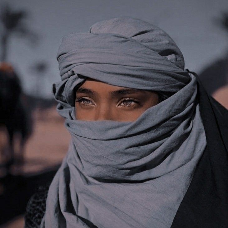 a woman wearing a gray head scarf and black jacket with her eyes wide open, standing in front of a horse