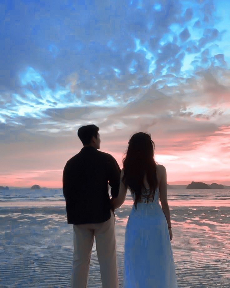 a man and woman standing on top of a beach next to the ocean at sunset