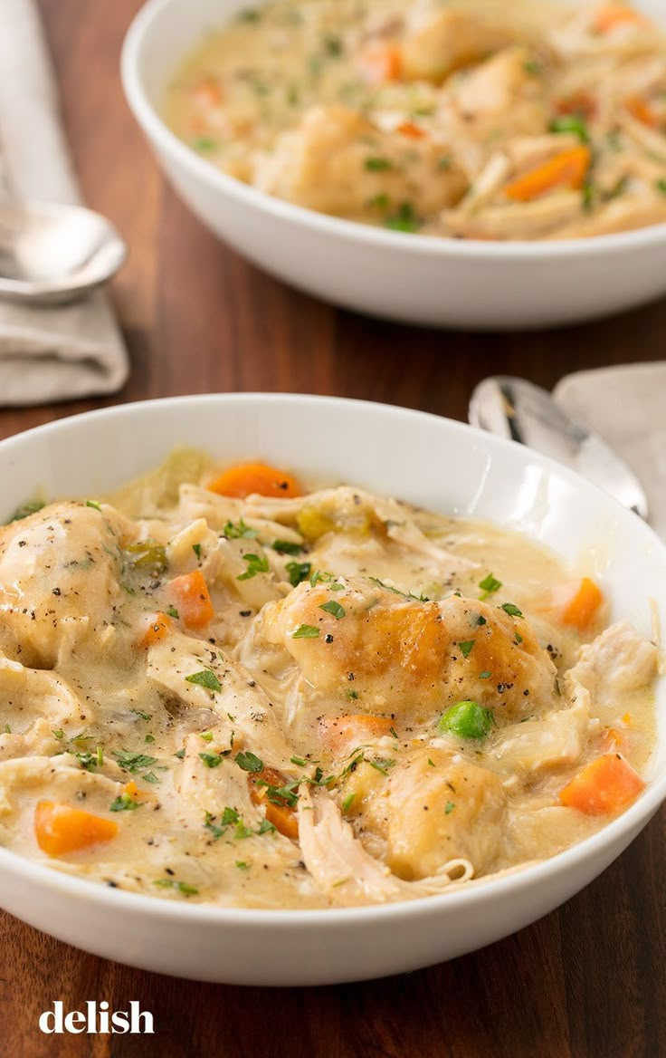 two bowls filled with chicken and dumplings on top of a wooden table next to silverware