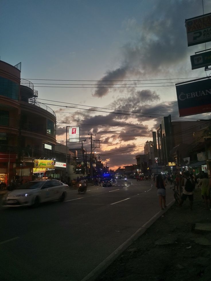 cars are driving down the street in front of tall buildings at dusk with people walking on the sidewalk