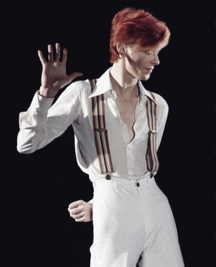 a man with red hair wearing white pants and suspenders is making a hand gesture