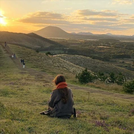 a woman sitting on top of a grass covered hillside next to a cow grazing in the distance